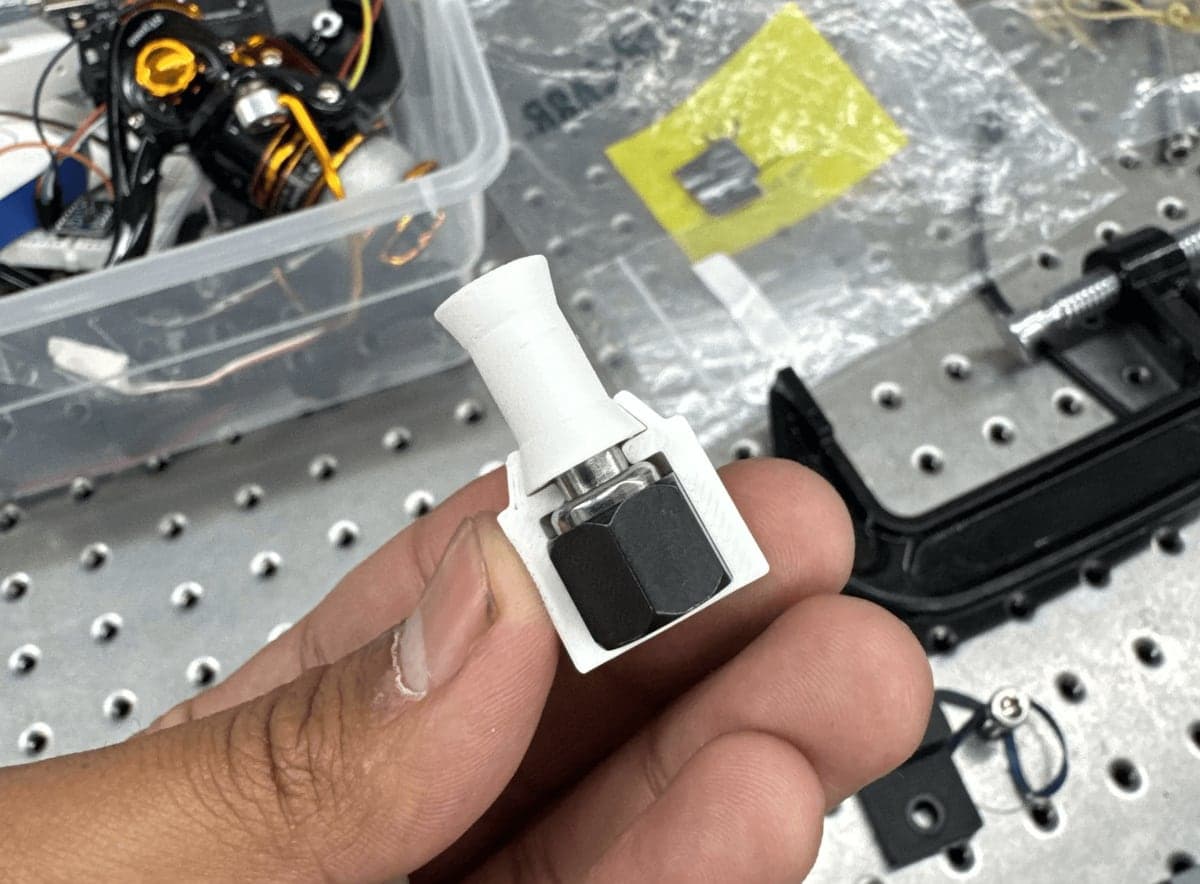 The compact white release mechanism held between finger and thumb above the test rig.