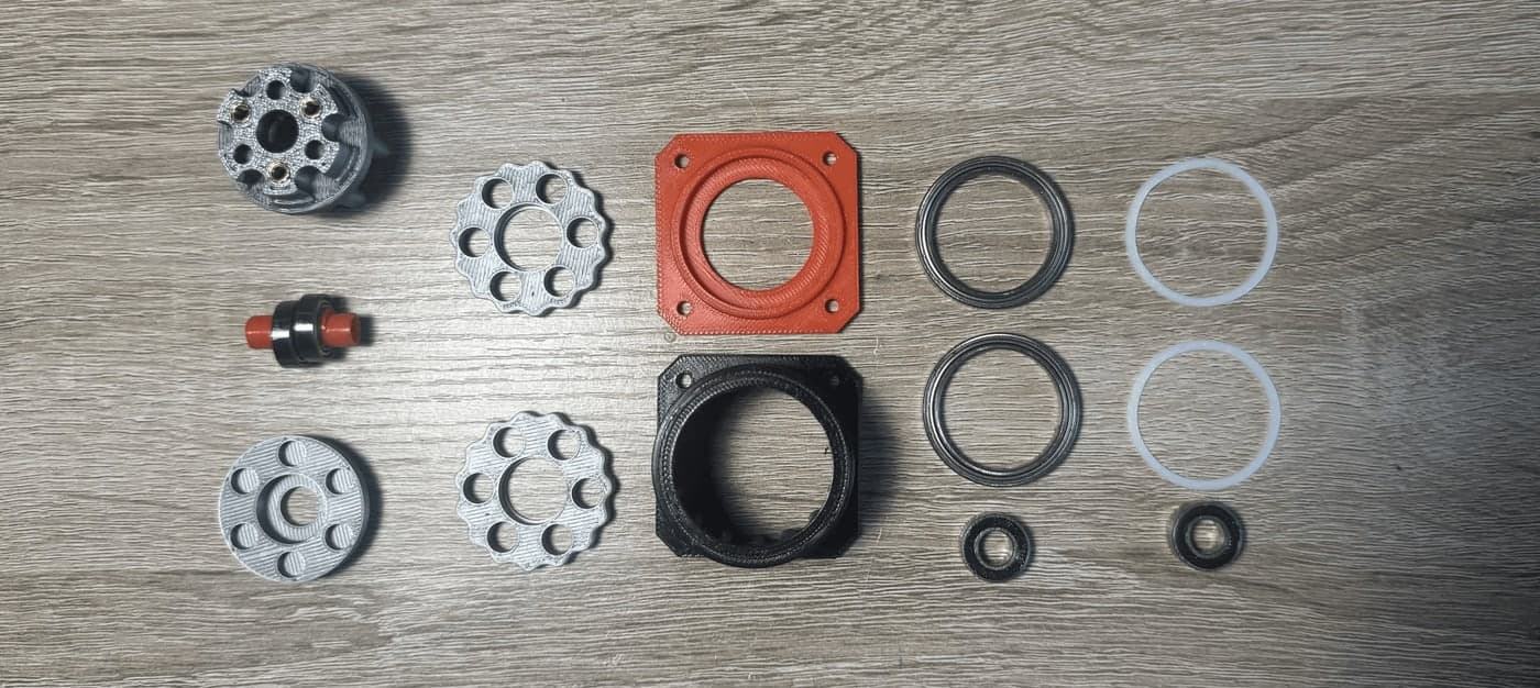 All gearbox components laid out on a workbench: output shaft, end cap, eccentric shaft, two cycloidal rings, black casing, orange closeout, bearings and PTFE washers.