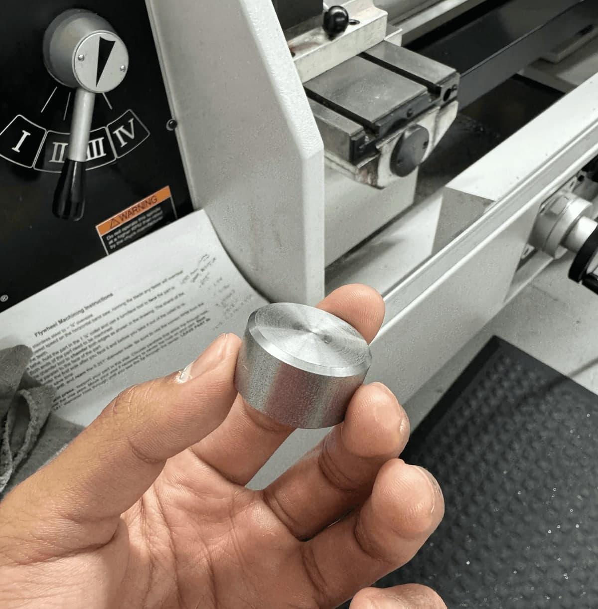 A freshly turned aluminium flywheel held in one hand in front of the lathe, with the flywheel machining instructions sheet resting on the machine behind it.
